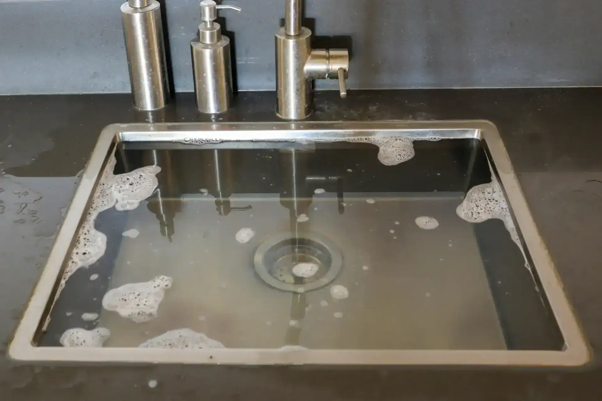 A stainless steel kitchen sink in FL is filled with soapy water, bubbles floating on the surface. Metal soap dispensers and a faucet sit on the countertop—perfect for a homeowner looking for a plumber Pasco County trusts.