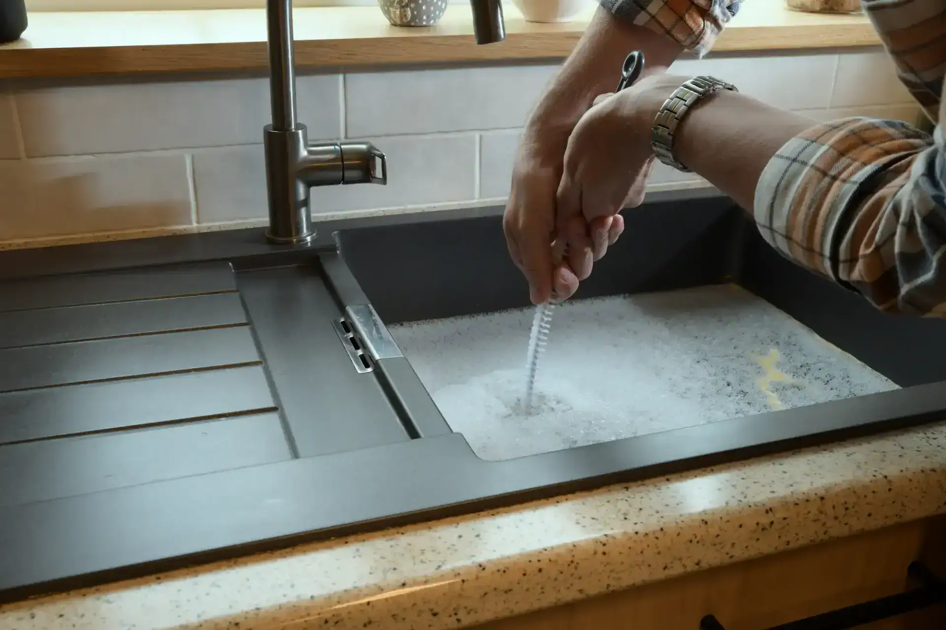 A person wearing a plaid shirt uses a brush to clean something in a kitchen sink filled with soapy water, next to a modern faucet and countertop, just like a professional plumber in Pasco County, FL might do.
