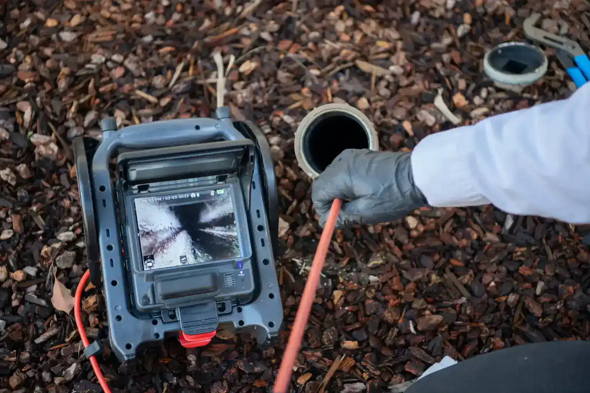 A person wearing a glove inserts a cable into an outdoor pipe while viewing the pipe’s interior on a video inspection monitor; the ground is covered with brown wood chips.